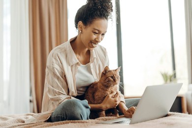 joyful african american young woman sitting on bed at home with her ginger cat, smiling while working on a laptop. online work concept