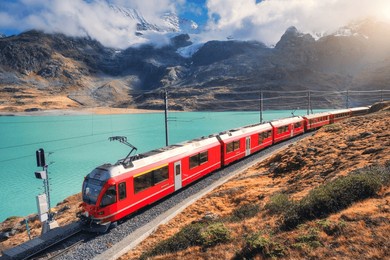 red train is moving along railway in swiss alps mountain range near lake on a sunny day with cloudy sky in autumn. landscape. bernina express train, orange grass, snowy rocks at sunset in fall. travel