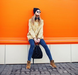 outdoor fashion portrait of stylish hipster cool girl wearing a black hat and sunglasses with backpack enjoying listens to music in the headphones against a colorful orange urban wall