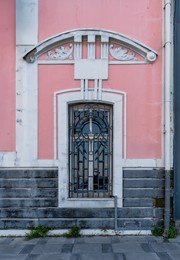 weathered pink building facade with ornate black metal window grate, white decorative architectural details, and grey stone base. urban street view