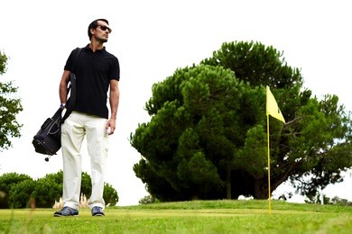 full length portrait of stylish golfer man in glasses standing on beautiful golf course looking away, handsome brunette hair man with golf bag on shoulder and hand in pocket standing on golf course