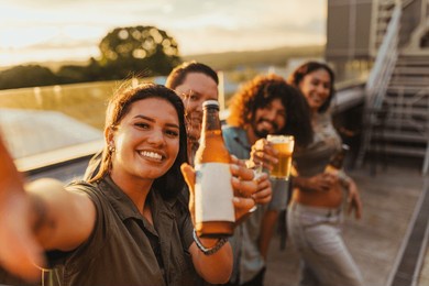 happy friends taking a selfie while toasting with beer on a rooftop at sunset