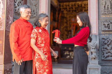 asian grandparents receiving red envelope from young woman at chinese temple, joyful moment of giving during lunar new year celebration, showing love, family connection and festive cultural tradition