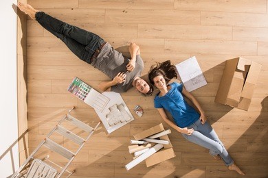 view from above, looking at the camera a young couple lying on the floor of their new home, there are blueprints to help them to think about their new interior decoration