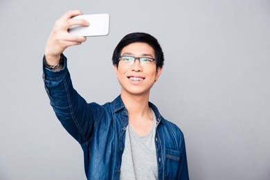 happy young asian man making selfie photo on smartphone over gray background