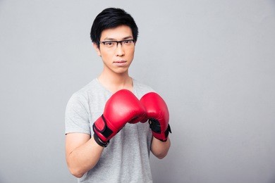 young asian man in glasses standing in boxing gloves over gray background
