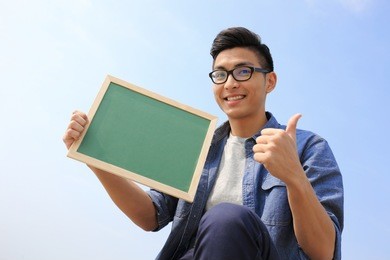 happy man holding blackboard or chalkboard and show thumb up with sky background, asian male