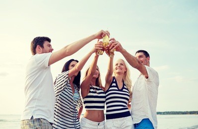summer, holidays, tourism, drinks and people concept - group of smiling friends clinking bottles of beer or cider on beach