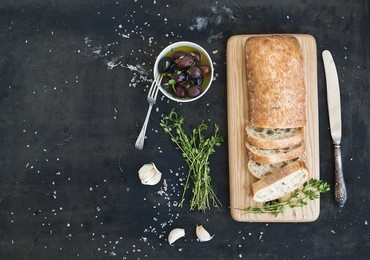 italian ciabatta bread cut in slices on wooden chopping board with herbs, garlic and olives over dark grunge backdrop, copy space, top view