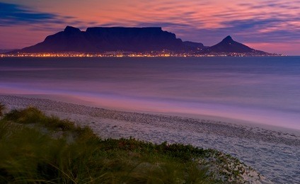 table mountain cape town landscape with lions head after sunset and city lights. ocean showing dimly lit beach front and peachy colored sky.