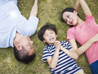 little asian boy lying on grass making a wish with eyes closed while his parents looking at him affectionately.