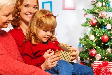 happy family with little daughter sitting on the laps of mother and grandmother opening little new year present with surprised face expression