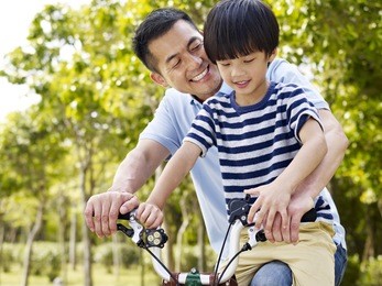 asian father and elementary-age son enjoying riding a bike outdoors in a park.