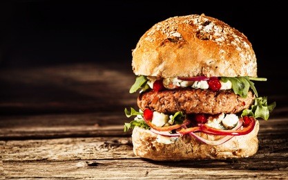 close up of burger piled high with fresh toppings on whole grain artisan bun, on rustic wooden surface with dark background and copy space