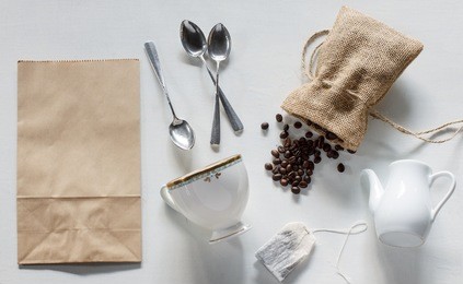 coffee beans in burlap bag, paper bag, silver spoon, vintage cup, white jar, teabag on white wood background.