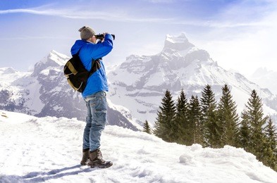 man with backpack is standing on a mountain and looking through binoculars.