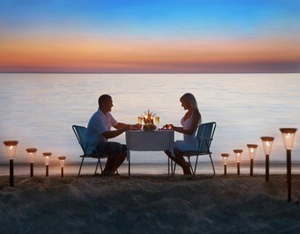 loving couple share a romantic dinner with candles, lanterns and wine glasses at sea beach sand against wonderful sunset