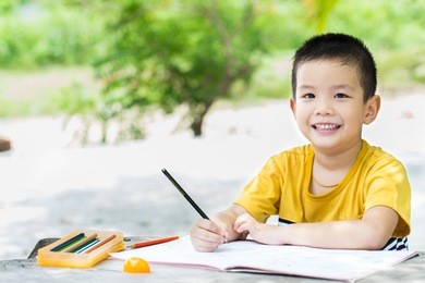 little asian boy use pencil writing on notebook for writing book with smiling face on wooden table in the park