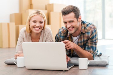 in search of a good moving company. cheerful young couple laying on the floor of their new apartment and looking at laptop while cardboard boxes laying in the background