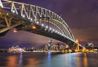 east side of sydney harbour bridge at sunset with bright illumination of steel arch and columns reflecting in the blurred waters of harbour with sydney city cbd in the background