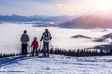 people observing mountain scenery.
family of three people stays in front of scenic landscape. these are skiers, they dressed in winter sport jackets and have skies attached.