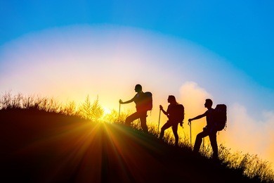 silhouettes of three people walking with backpacks and other hiking gear up toward top of wild grass mountain mother father daughter bright luminous sunrise sky background 