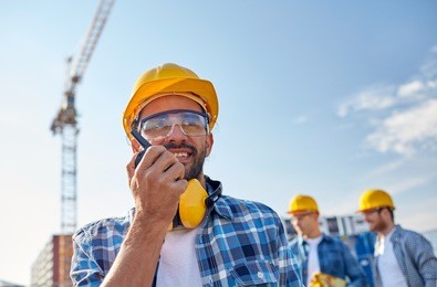 industry, building, technology and people concept -male builder in hardhat with walkie talkie or radio at construction site