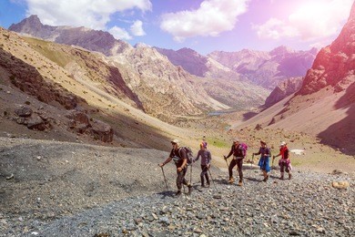 diverse group of hikers in mountain terrain.
people walking along rocky footpath positive faces trekking gear poles sporty clothing bright panorama blue sky sunny day background
