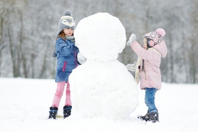 two funny adorable little sisters building a snowman together in beautiful winter park during snowfall