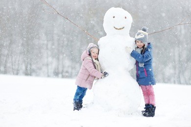 two funny adorable little sisters building a snowman together in beautiful winter park during snowfall