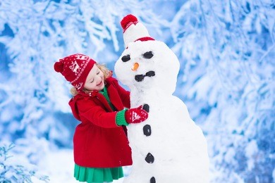 funny little toddler girl in a red knitted nordic hat and warm coat playing with a snow man. kids play outdoors in winter. children having fun at christmas time. child building snowman at xmas.