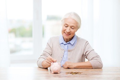 savings, money, annuity insurance, retirement and people concept - smiling senior woman putting coins into piggy bank at home