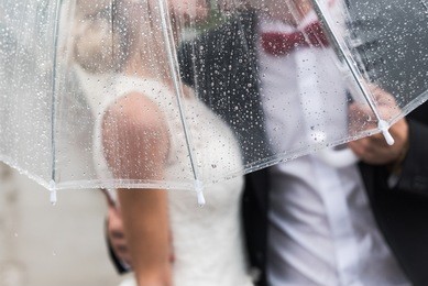 the bride and groom in the rain are covered with a transparent umbrella, rain drops