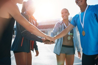 shot of a group of young sportsmen with medals piling their hands while standing in a huddle. successful team of athletes with their hands together cheering victory.