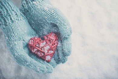 woman hands in light teal knitted mittens are holding a beautiful glossy red heart in a snow winter background. love and st. valentine concept.