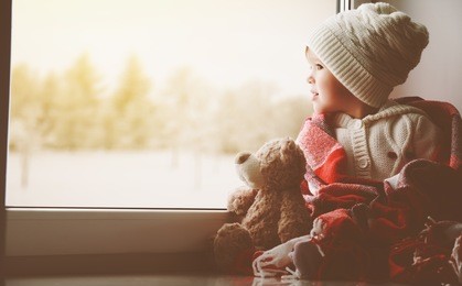 child little girl sitting by the window with a teddy bear and looking at the winter forest