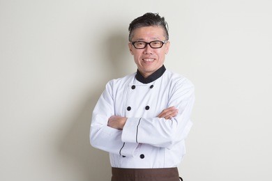 portrait of confident 50s mature asian male chef in uniform arms crossed, standing on plain background with shadow, copy space.