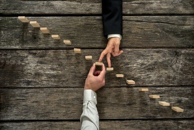 conceptual image of business partnership and support - businessman supporting wooden step in a staircase made of pegs as his partner walks his fingers up towards growth, achievement and development.