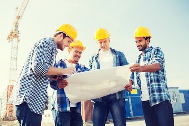 business, building, teamwork and people concept - group of smiling builders in hardhats with clipboard and blueprint outdoors