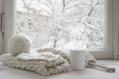cozy winter still life: mug of hot tea and warm woolen knitting on vintage windowsill against snow landscape from outside.