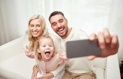holidays, technology and people concept - happy family sitting on sofa and taking selfie picture with smartphone at home