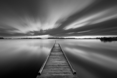 a small jetty on a lake near amsterdam the netherlands in black and white. a slow shutter speed was used to see the movement of the clouds in the sky. photographed at dawn.