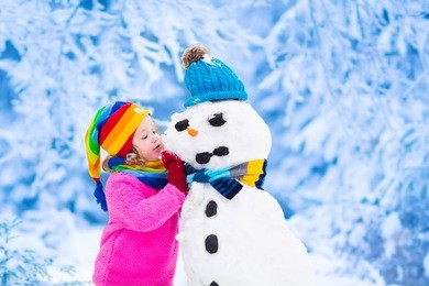 funny little toddler girl in a colorful hat and warm coat playing with a snow man. kids play outdoors in winter. children having fun at christmas time. child building snowman at xmas.
