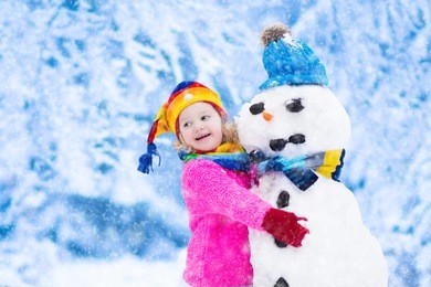 funny little toddler girl in a colorful hat and warm coat playing with a snow man. kids play outdoors in winter. children having fun at christmas time. child building snowman at xmas.
