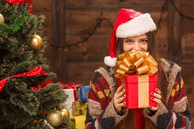 beautiful girl holding a christmas present in front of her. happy woman in santa hat standing near new year tree and waiting for celebration.