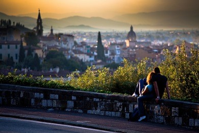 a couple in love - girl and boy sitting on a small wall by the road watching a scenic sunset over a romantic italian city on the hills in the blurred background; in florence, italy