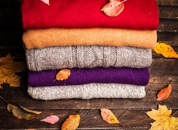 pile of knitted winter clothes on wooden background covered with autumn leaves, knitwear, space for text. stack of knitted sweaters and cardigans.