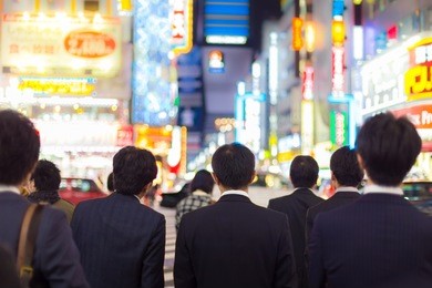 japanese corporative business people in suits, waiting in rush hour on crossroad in shinjuku business district, tokyo, japan. blured advertising boards illuminated in the background.