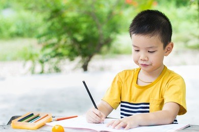 little asian boy use pencil writing on notebook for writing book with smiling face on wooden table in the park