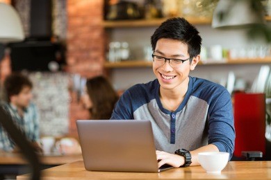 happy cheerful young asian male in glasses smiling and using laptop in cafe
